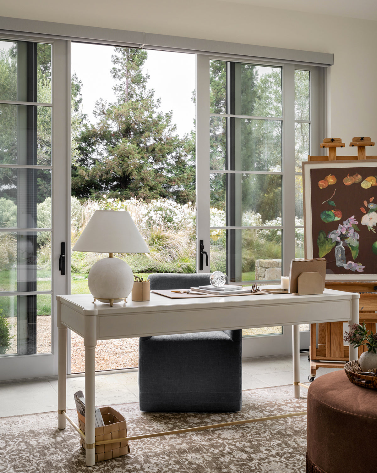 A bright, elegant workspace with a white desk and chair features the Price Table Lamp, its alabaster base and linen shade beside large glass doors. An easel displays a floral painting as sunlight fills the room overlooking a garden, by McGee & Co.