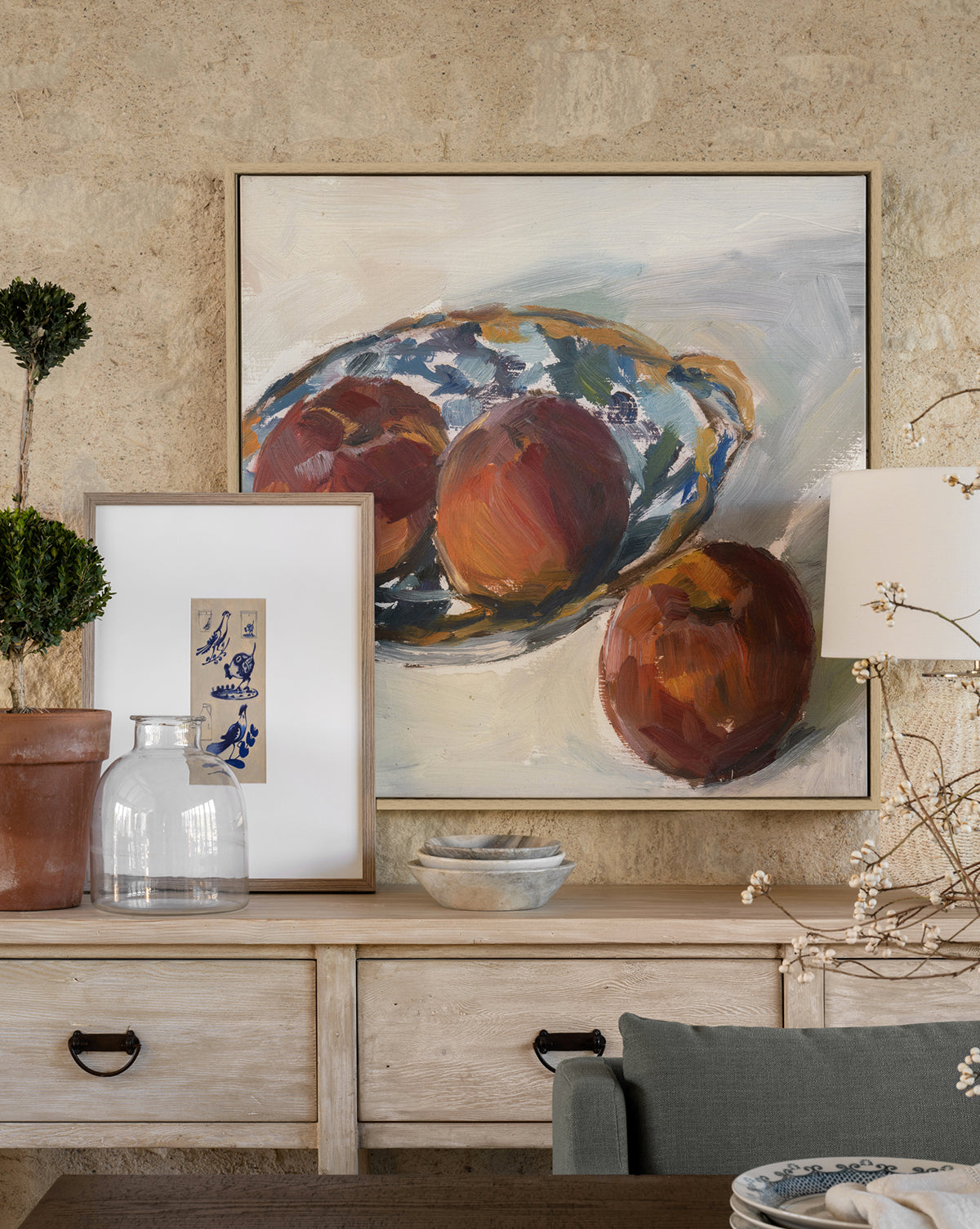 A framed painting of three apples on a blue and white plate hangs above a wooden console table decorated with framed art, a potted plant, a Kelby Glass Vase, a bowl, and a lamp with white flowers.