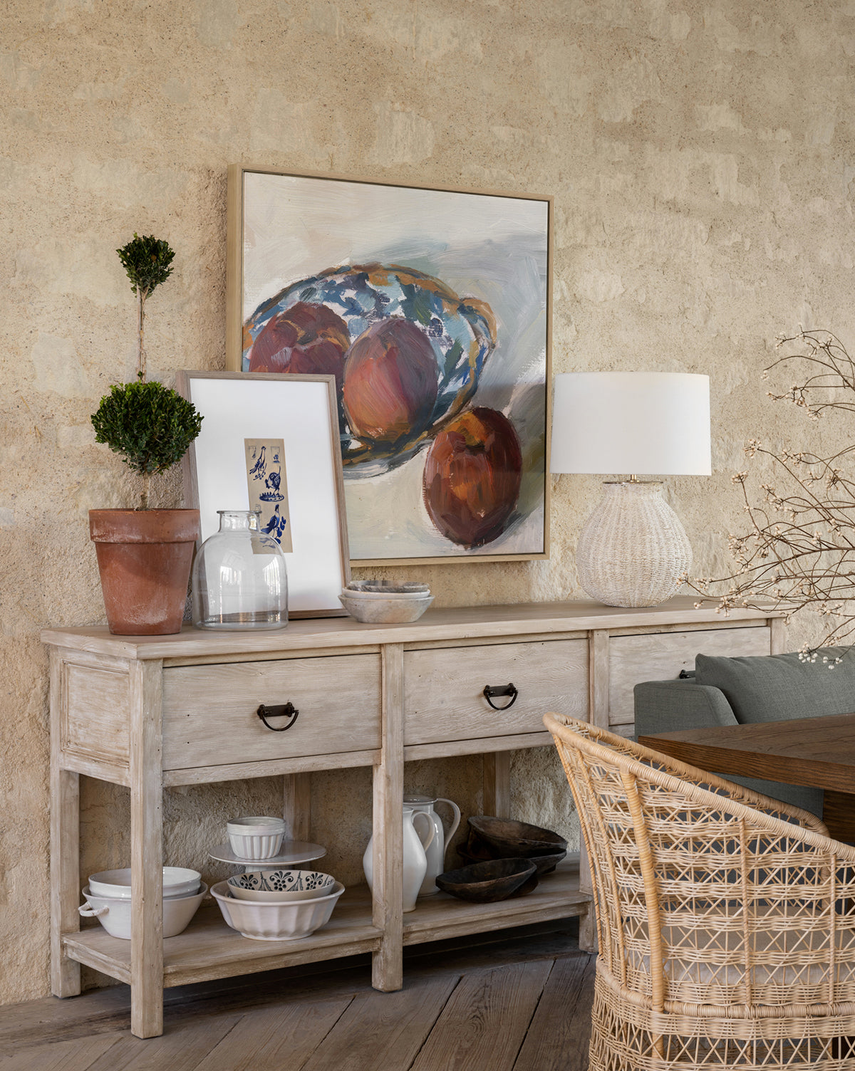 The Martha Sideboard, made from reclaimed Douglas Fir, showcases potted plants, art frames, a glass pitcher, bowl, and lamp against a stone wall. A wicker chair and dining table sit nearby, by McGee & Co.