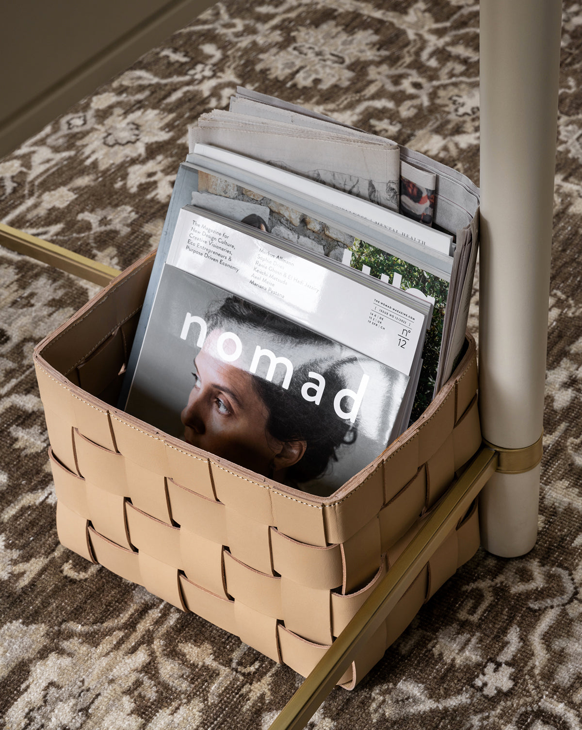 The Rupert Leather Bin sits on a patterned rug, holding magazines and newspapers—ideal for a stylish home office. On top is the magazine nomad, its cover showing a close-up of a person’s face.