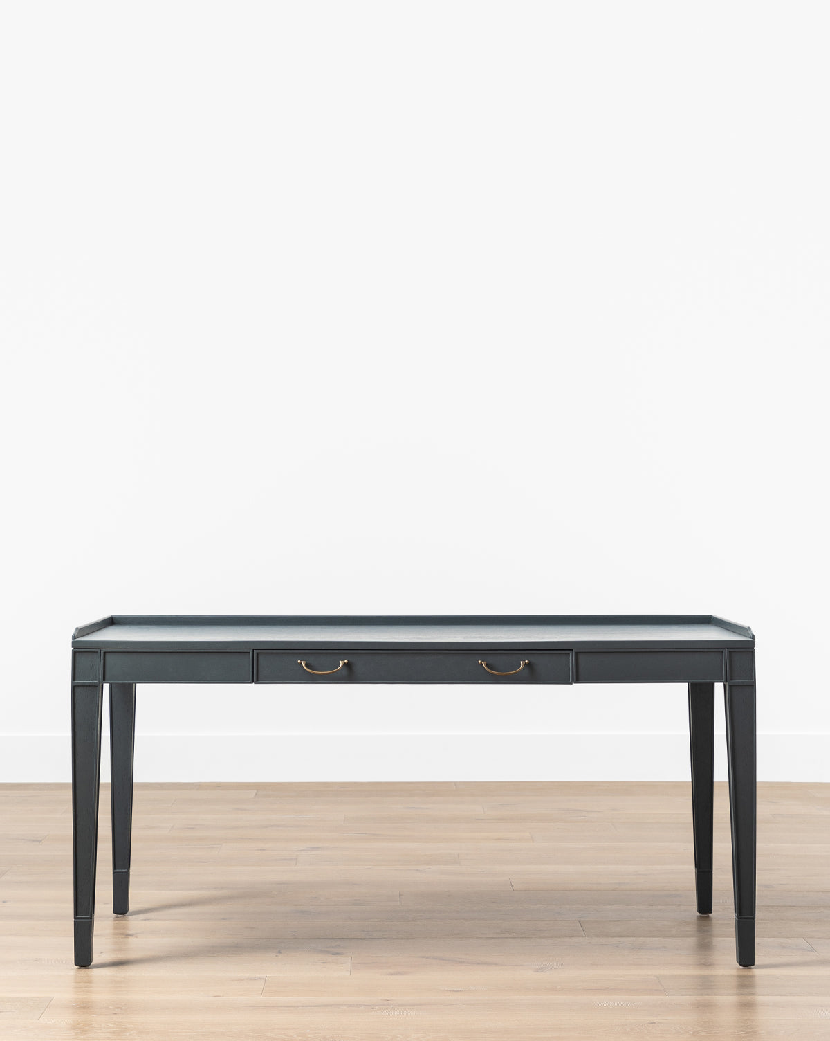 The Stratford Desk is a black rectangular home office desk with a dark slate matte top, slender legs, and two central drawers with brass handles, shown on a light wooden floor against a plain white wall by McGee & Co.