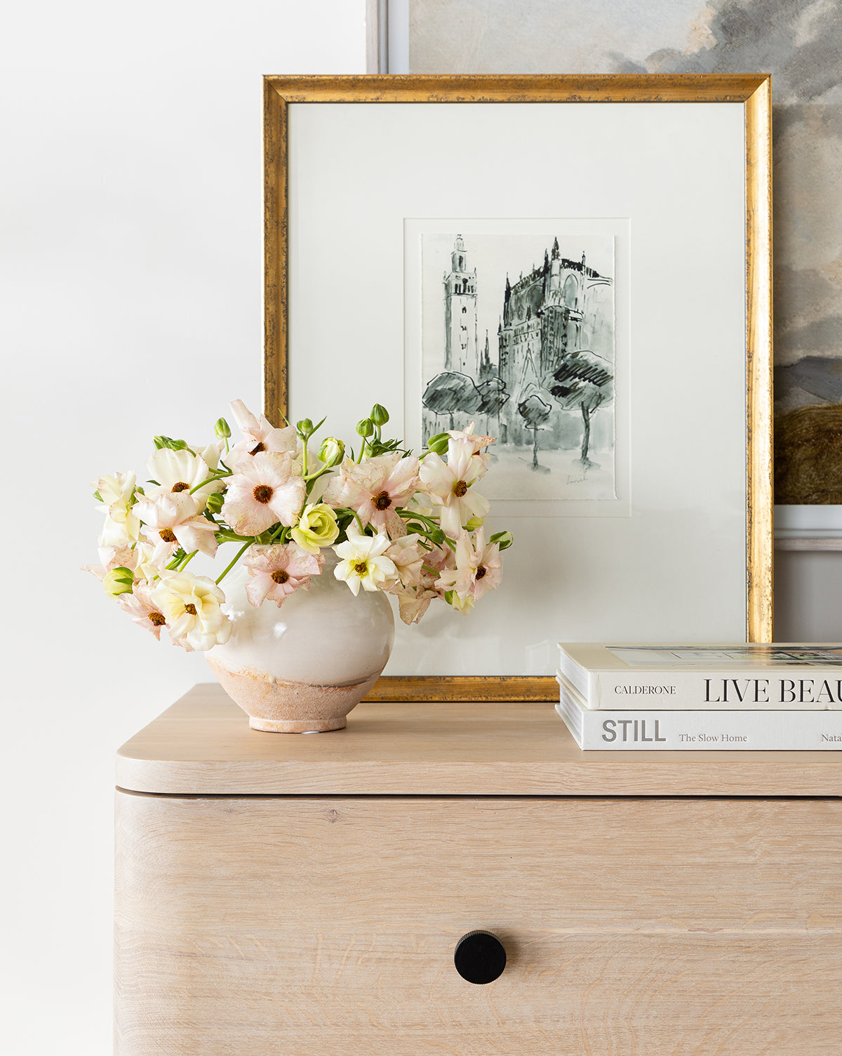 A light wood dresser topped with stacked books—including “LIVE BEAUTIFULLY”—a small round vase of pale pink and white flowers, and a gold-framed Castle Sketch arranged neatly.