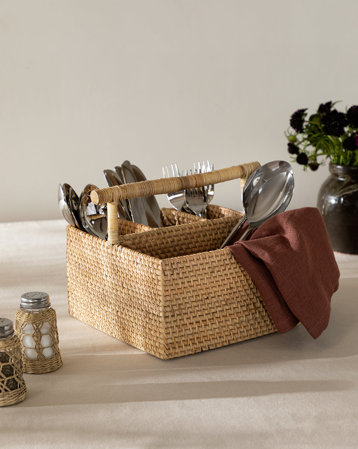 The Rattan Utensil Caddy with a handle stores silverware and a brown napkin. Salt and pepper shakers are nearby, and a dark vase with flowers sits in the background on a beige tablecloth, by McGee & Co.