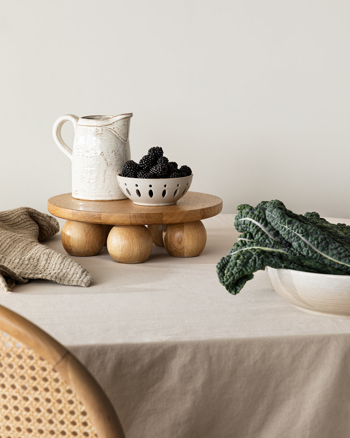 A White Stoneware Pitcher sits on a round wooden stand atop a beige tablecloth, next to a bowl of blackberries, leafy greens, and a nearby wicker chair, by McGee & Co.