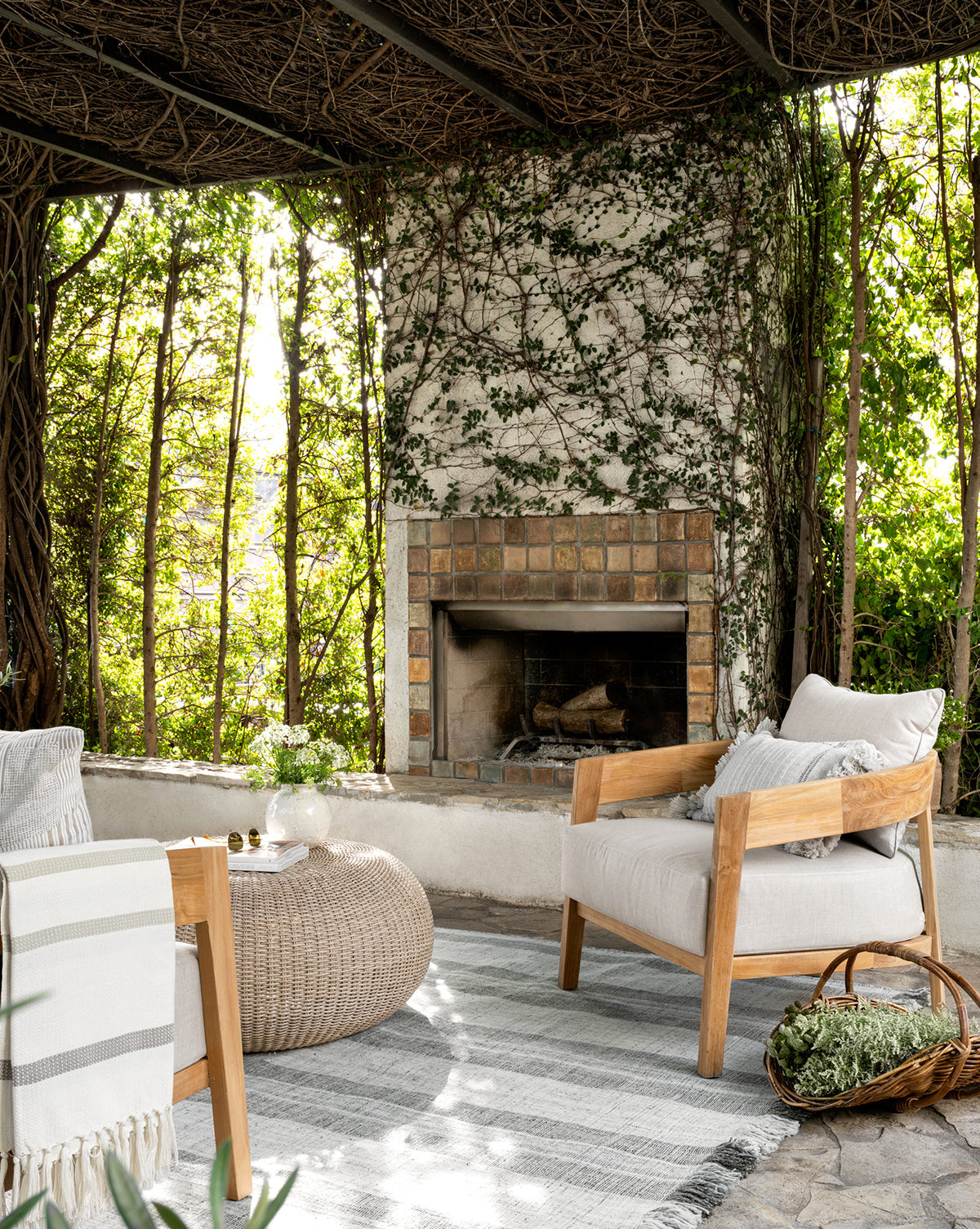 A cozy patio features Vento Teak Outdoor Lounge Chairs, a woven ottoman, striped rug, and an ivy-clad fireplace. Sunlight streams through greenery and latticework, creating a serene and inviting outdoor retreat by McGee & Co.