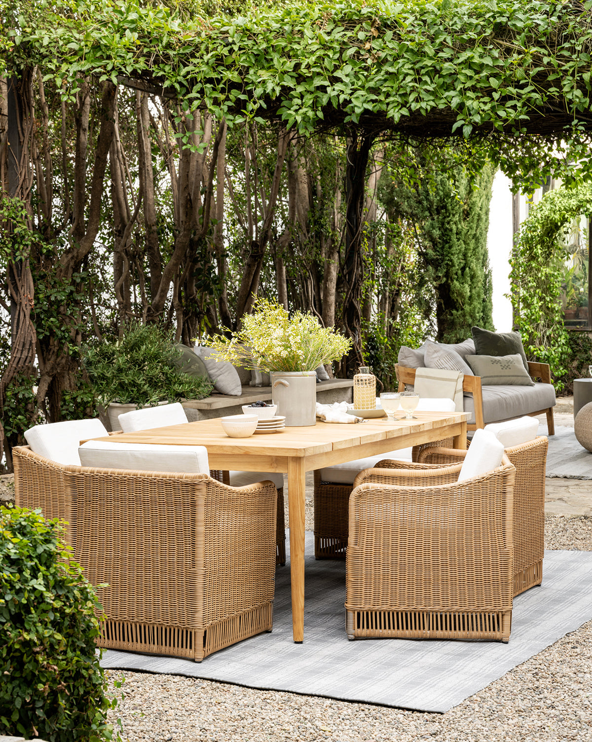 Outdoor patio featuring the Elowyn Outdoor Dining Table, six wicker chairs with white cushions, a gray outdoor sofa, and lush greenery overhead for a shaded, welcoming garden dining experience in any weather, by McGee & Co.