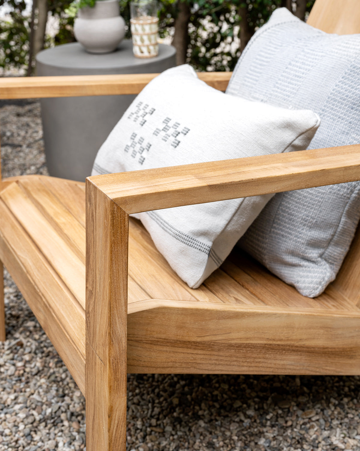 A close-up of the Triby Teak Outdoor Lounge Chair with two textured light gray pillows sits on a gravel patio beside a round gray side table holding a vase and glass, with green foliage in the background, by McGee & Co.