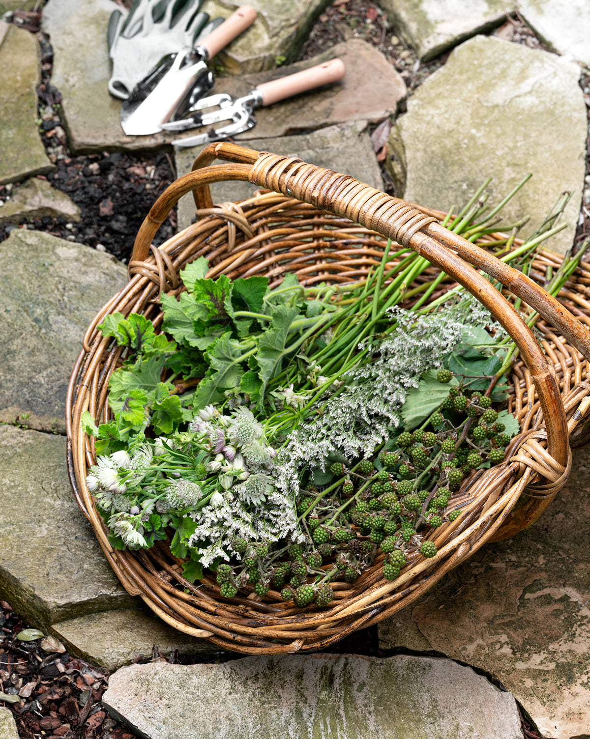 A Rattan Gardening Tray holds freshly picked green herbs and plants on stone paving, accented by gardening tools with wooden handles for an organic, natural touch in a peaceful outdoor setting.