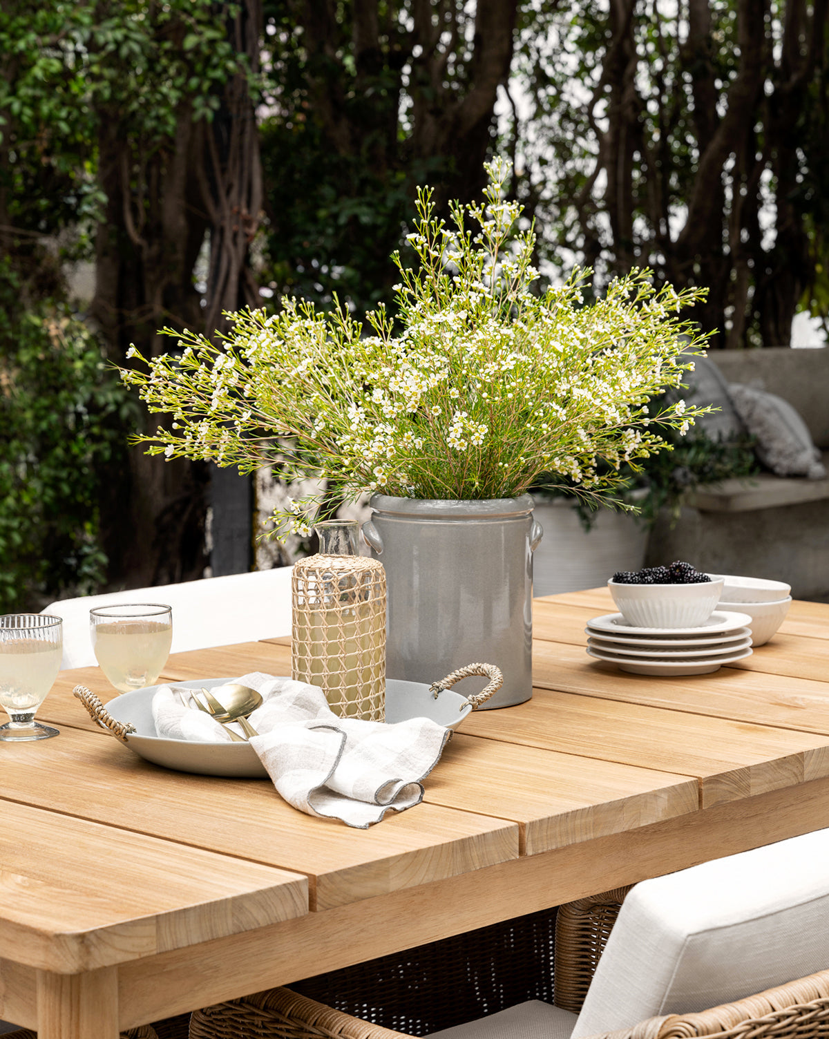 The Elowyn Outdoor Dining Table is set with white plates, bowls of blackberries, glasses of lemonade, napkins, and a gray ceramic vase with white wildflowers. Cushioned chairs and lush greenery complete the inviting outdoor scene by McGee & Co.