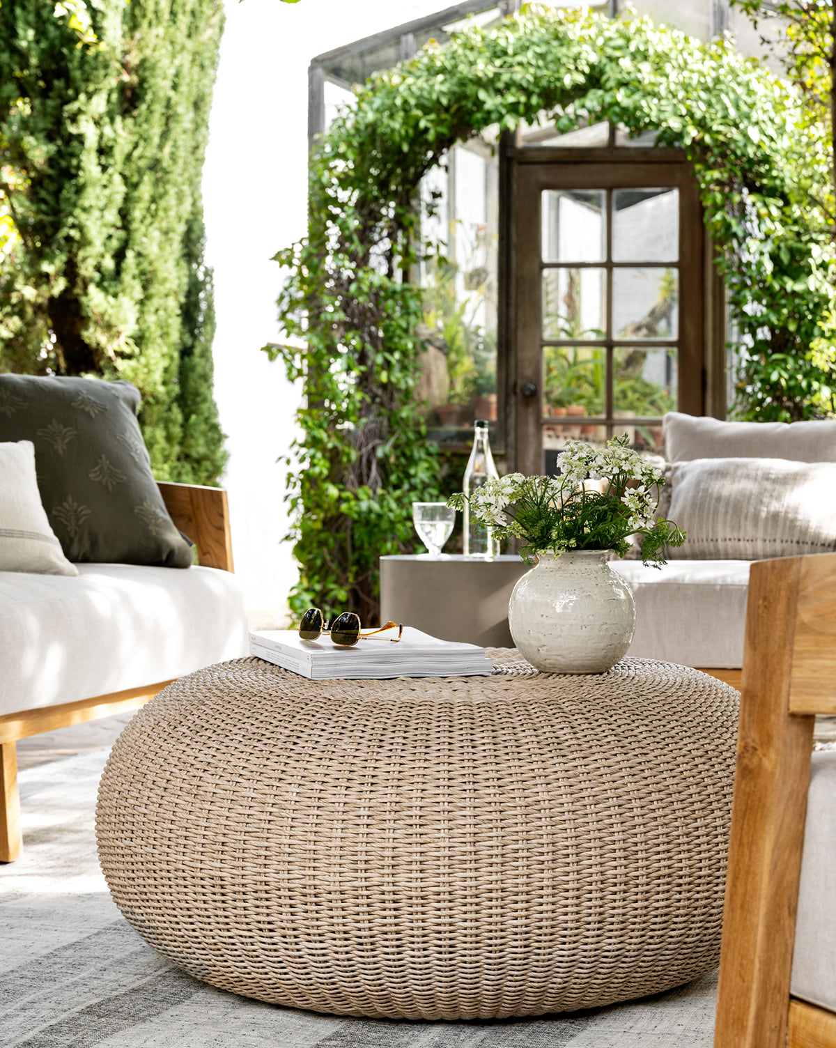 A cozy patio features beige cushioned chairs and the Rimma Outdoor Coffee Table, crafted from all-weather wicker, topped with a white vase of flowers and a book. Lush green ivy arches and a glass door frame the sunlit garden beyond, by McGee & Co.