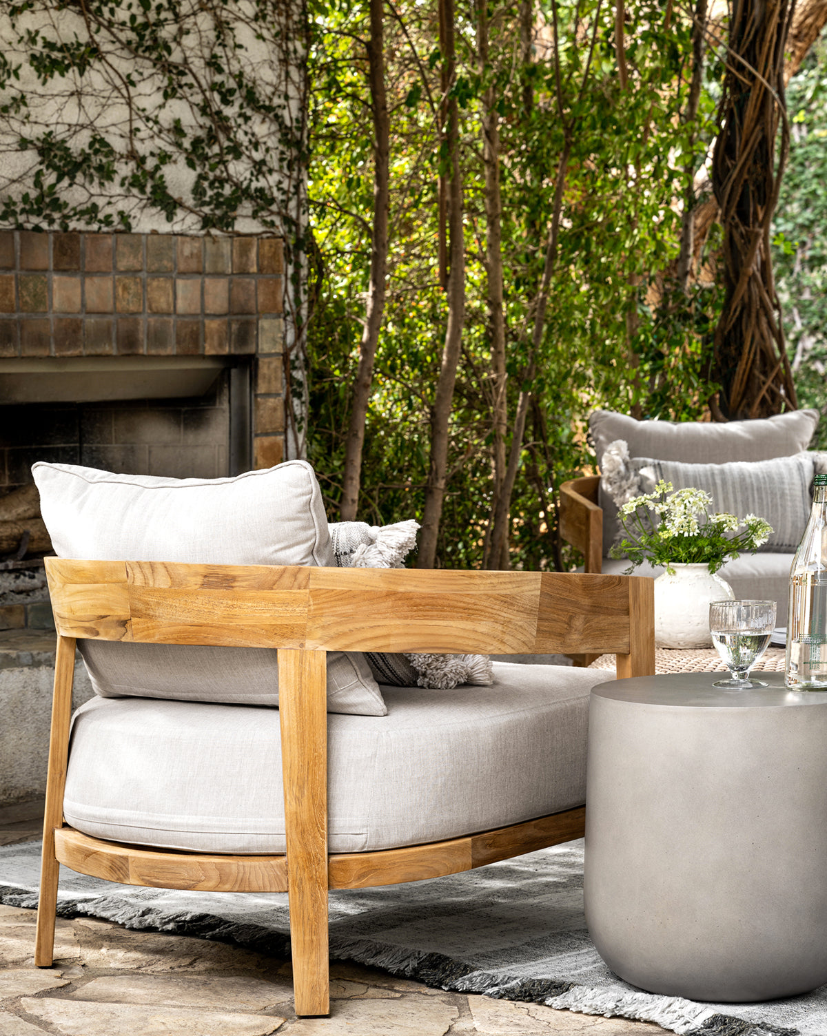 A cozy outdoor patio features the Vento Teak Outdoor Lounge Chair, a round gray side table with glassware, and green plants in the background, creating a relaxing, natural setting by McGee & Co.