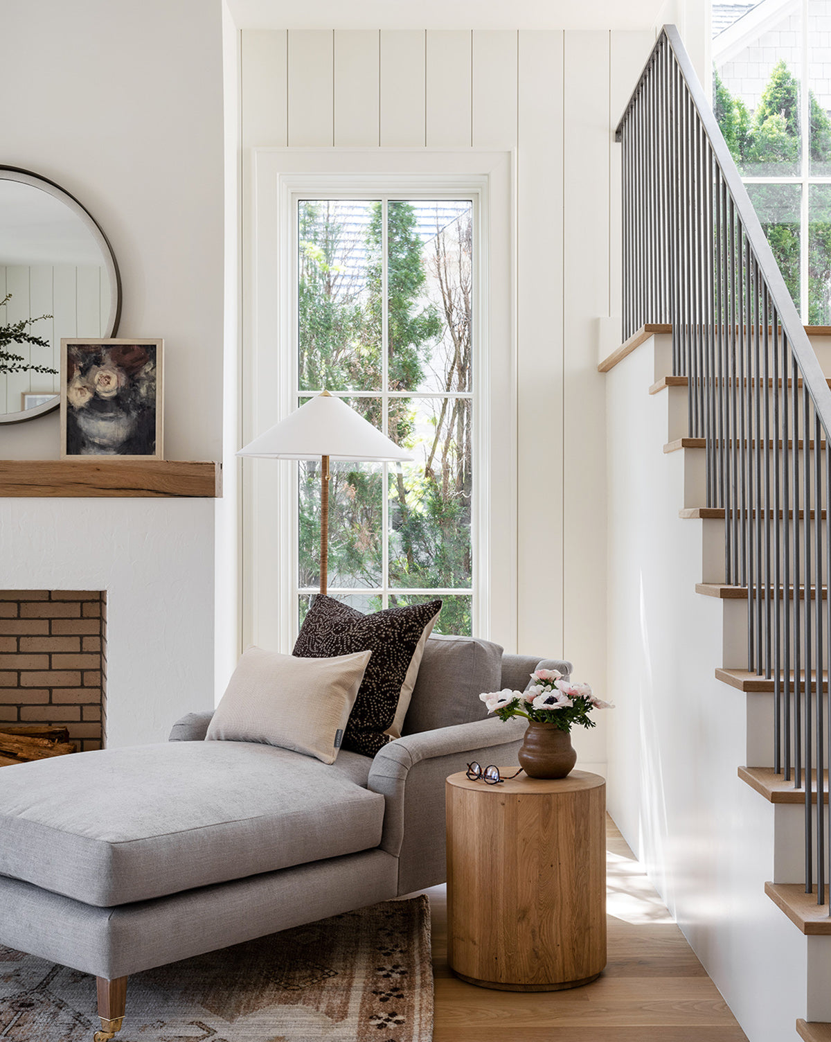 A bright, modern living room with a gray chaise lounge and neutral pillows showcases a round wooden side table topped with Midnight Bouquet by Leslie Dayas in a vase, plus a large window, fireplace mirror, and staircase with black railings.