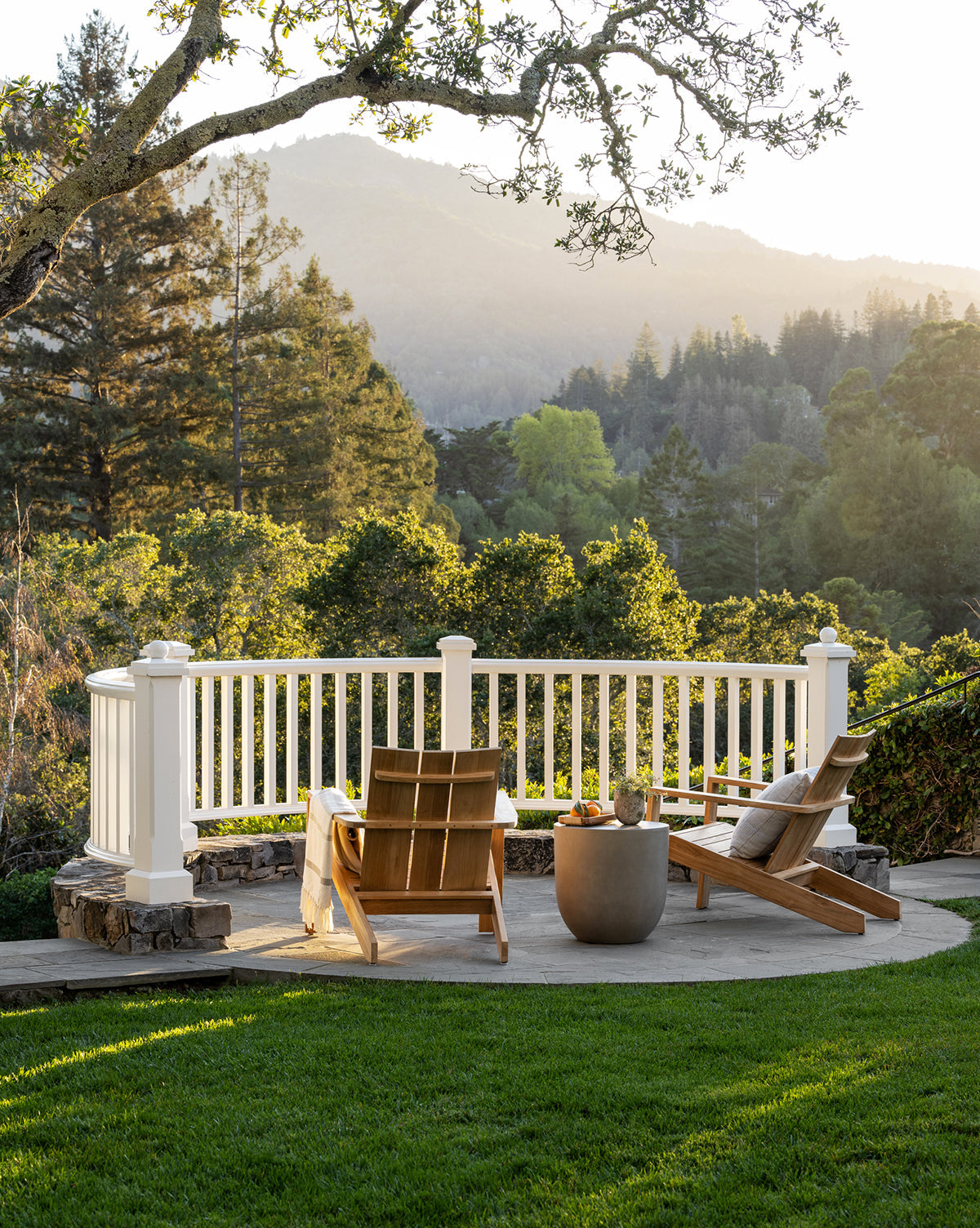 Two Triby Teak Outdoor Lounge Chairs sit on a patio facing a white railing, separated by a small table. Trees and distant mountains are softly illuminated by sunlight beyond this all-weather arrangement, by McGee & Co.