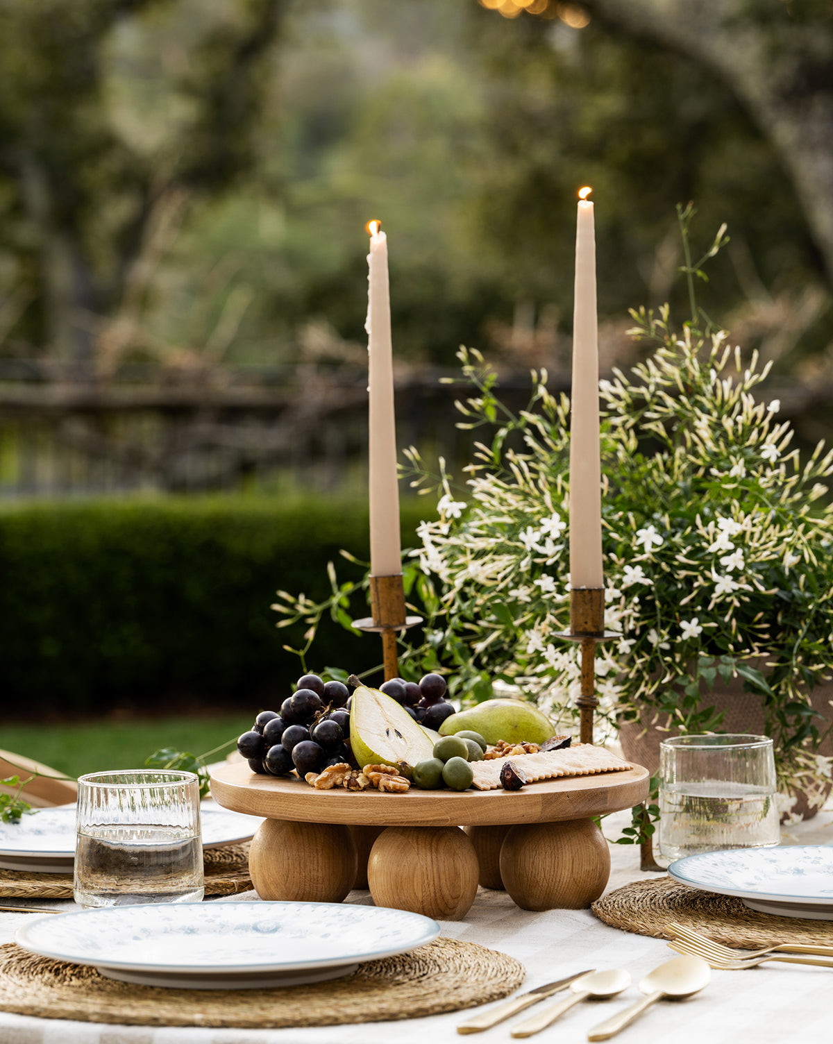 A rustic outdoor table is set with two lit candles, a Bronx Oak Pedestal displaying assorted fruits, cheese, and nuts, two place settings with glassware, and greenery in the background by McGee & Co.