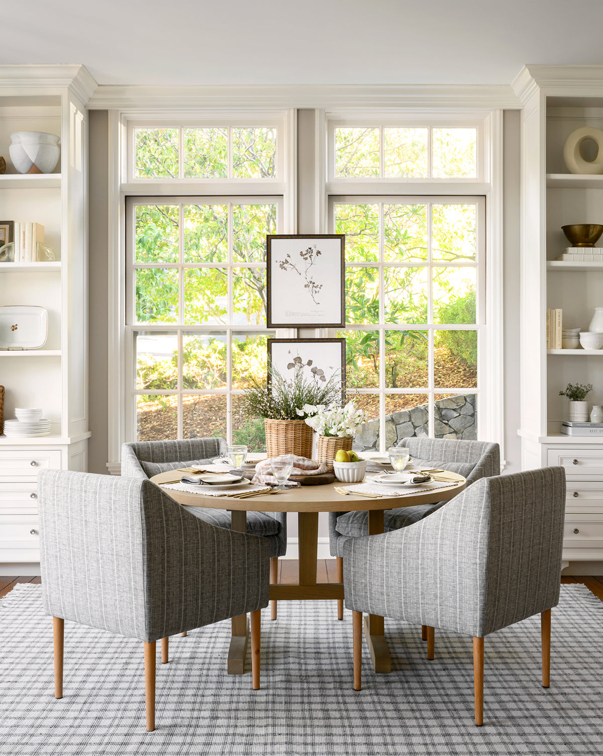 A bright dining area with large windows showcases a Giselle Round Table in white oak set for four, gray upholstered chairs, a white floral centerpiece, and decorated shelves, with greenery visible through the windows by McGee & Co.
