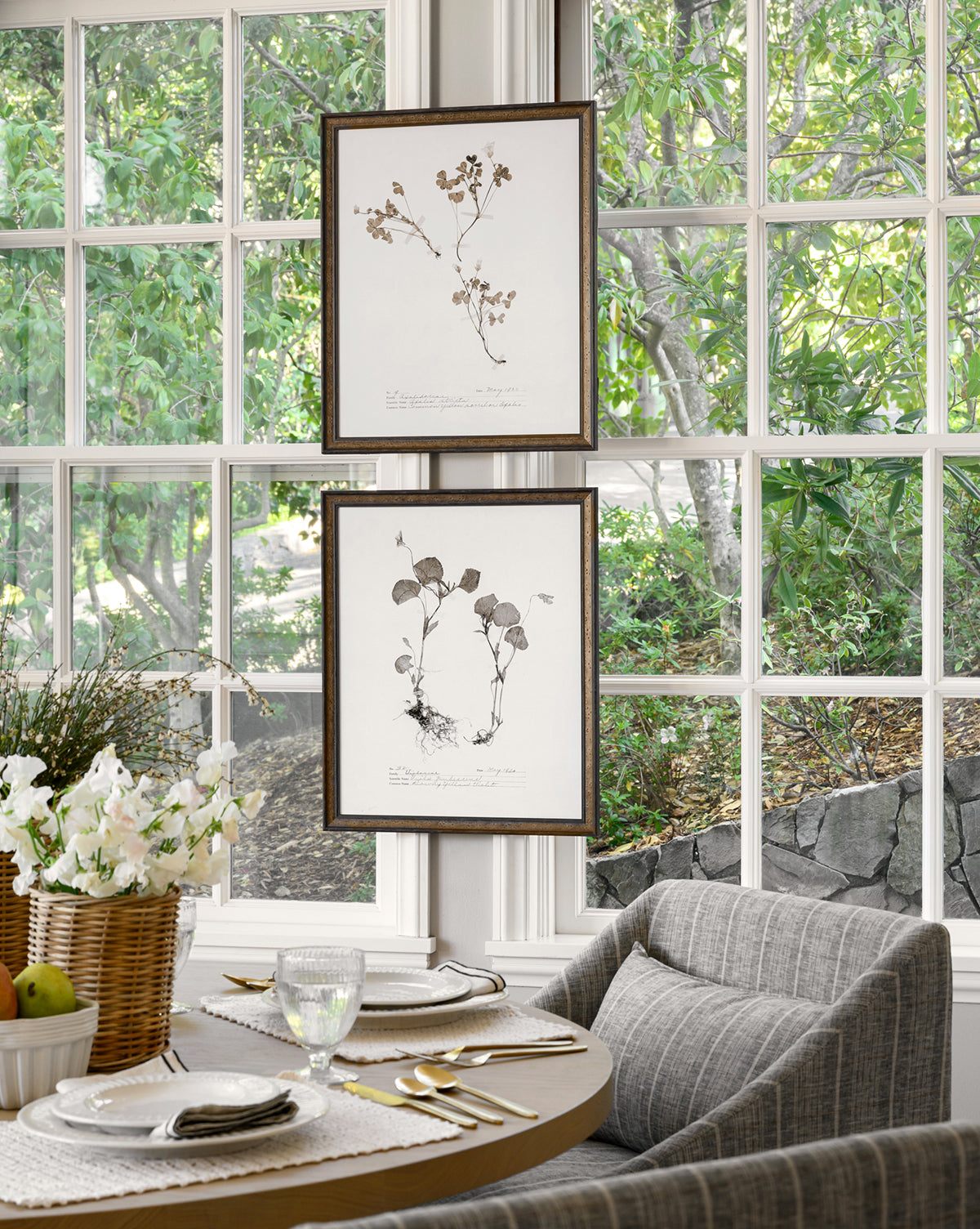 A cozy dining nook features a round table for four, gray cushioned chairs, a basket of white flowers, and two Subdued Botanicals I framed prints hanging before large windows with leafy green views.