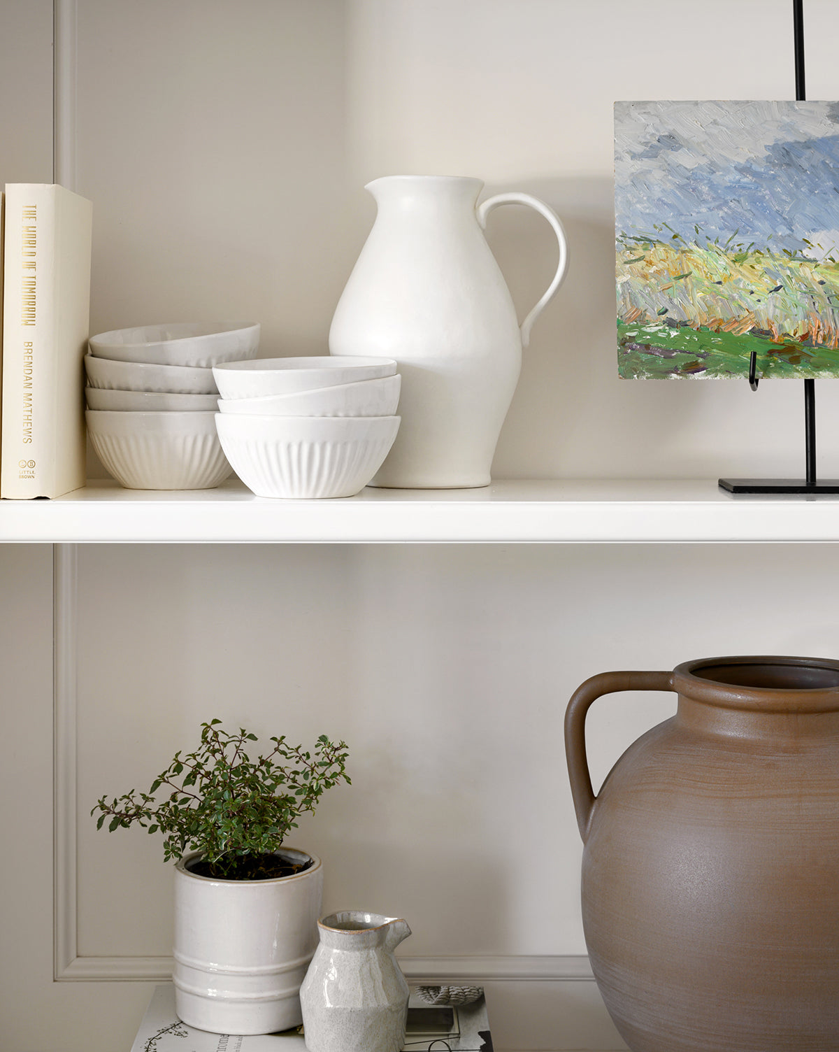 Two white shelves showcase stacked Adele Bowls with fluted detailing, a white pitcher, a small canvas painting on a stand, a potted plant, a grayish jug, and a large brown pot. A book stands upright at the left end, by McGee & Co.