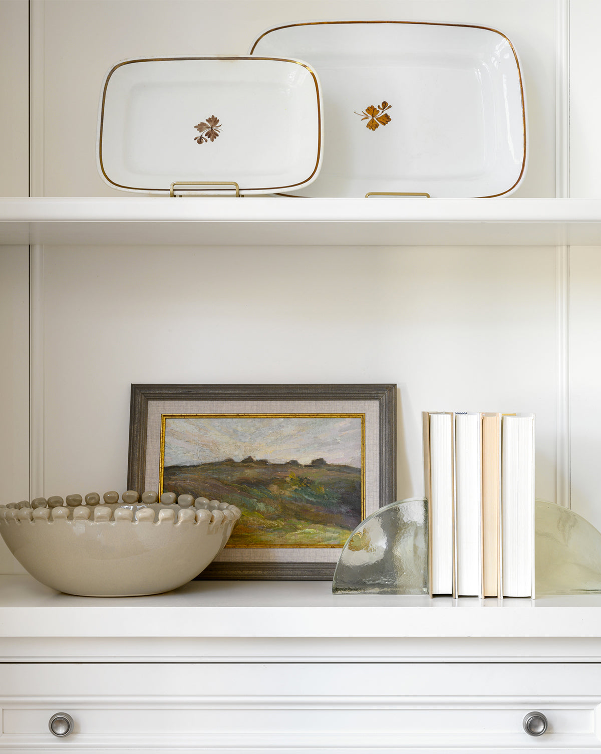 A white shelf holds two decorative plates, the Malibu Bowl, a framed landscape painting, books in a row, and two glass bookends. The all-white background and furniture give a clean look ideal for showcasing unique kitchen décor by McGee & Co.