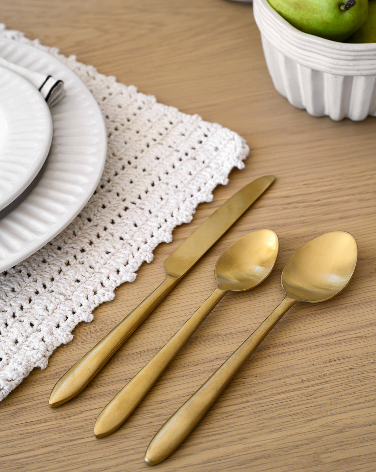 The gold Lilia Flatware (Set of 5) is displayed on a light wooden table alongside a white crocheted placemat, stacked white plates, and a white bowl filled with green pears.