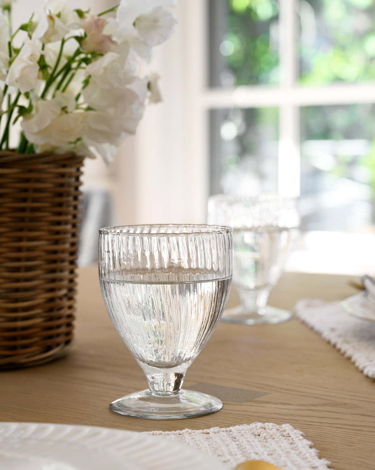 A Ribbed Wine Glasses (Set of 4) sits on a wooden table, filled with water, beside white flowers in a wicker basket as sunlight streams through the window in the background, by McGee & Co.