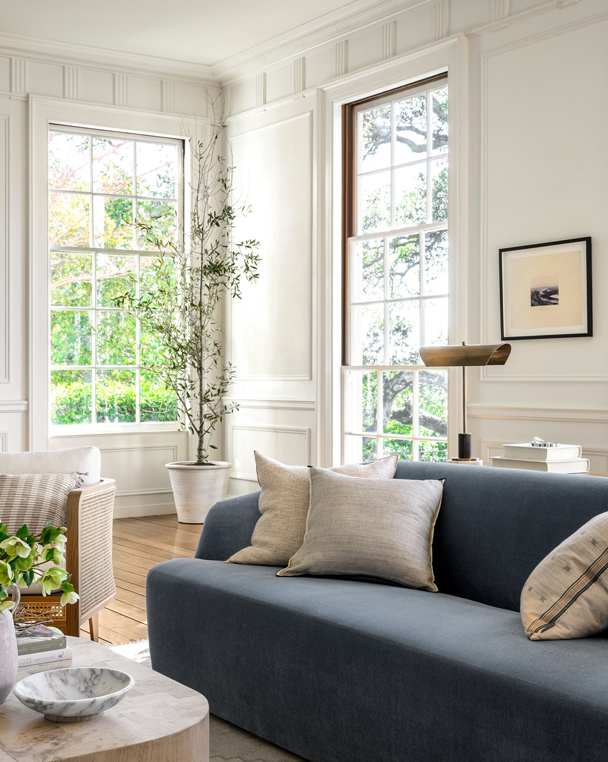 A bright, elegant living room with large windows, white paneled walls, a dark gray sofa, minimal art, and natural light—featuring the museum-quality framed art piece Palos Verdes.