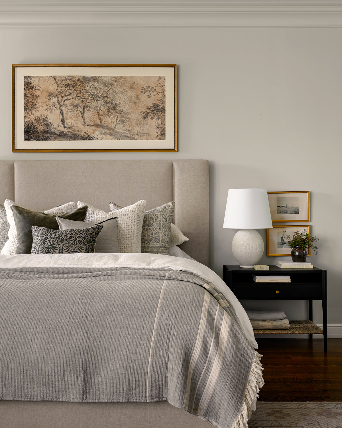 A neatly made bed with neutral bedding and pillows sits by a black nightstand with a white lamp, art, and plant. Above the bed hangs the Dutch Landscape Study artwork in an antique gold frame.