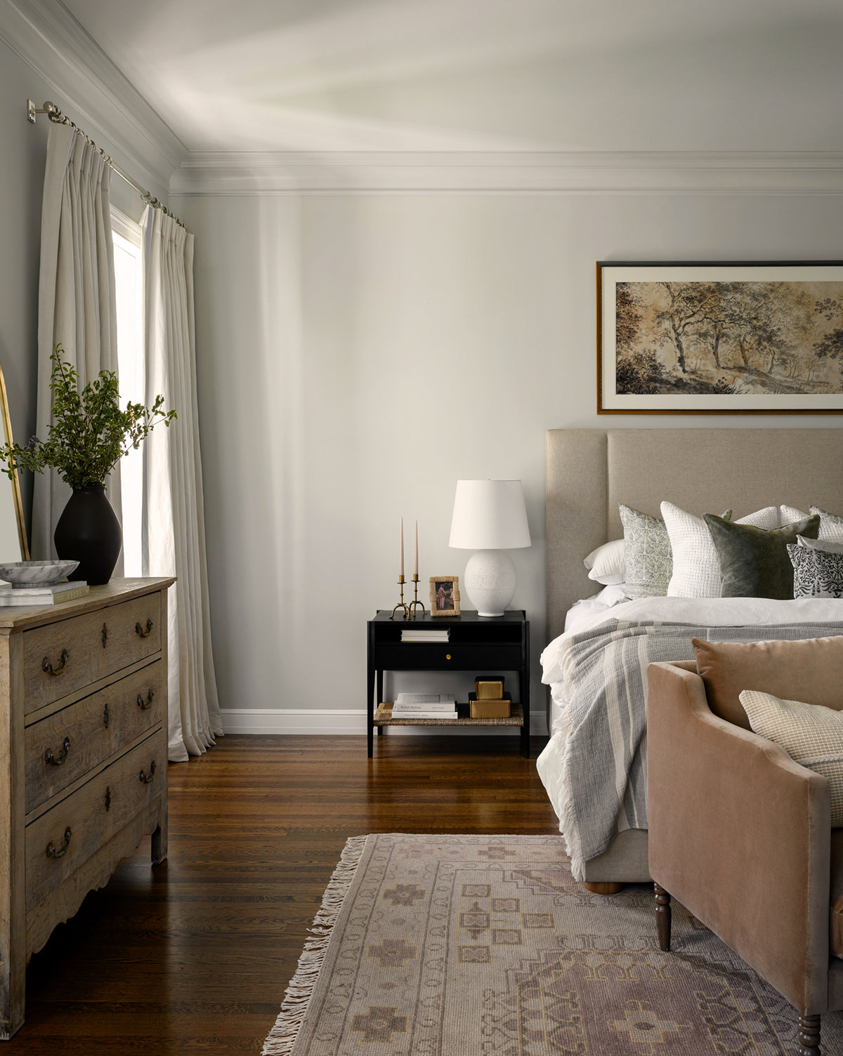 A cozy bedroom features a beige upholstered bed with layered pillows, a wooden nightstand and lamp, a vintage dresser with greenery, hardwood floors, and the Kenna Hand-Knotted Wool Rug—a perfect complement to the large landscape painting, by McGee & Co.