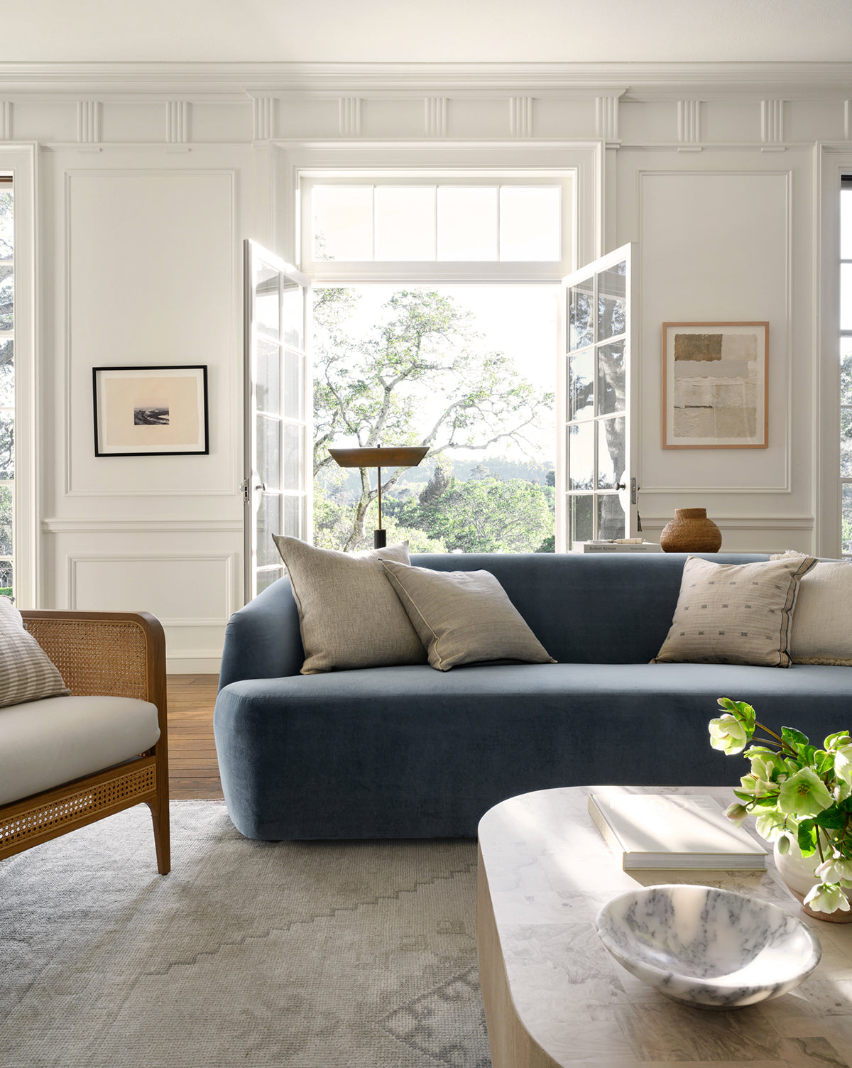 A bright, elegant living room features the Makerspalm Byron Sofa 96 with light cushions, a wooden armchair, art on white walls, large French doors opening to greenery, and a coffee table with a bowl, book, and flowers—styled by McGee & Co.