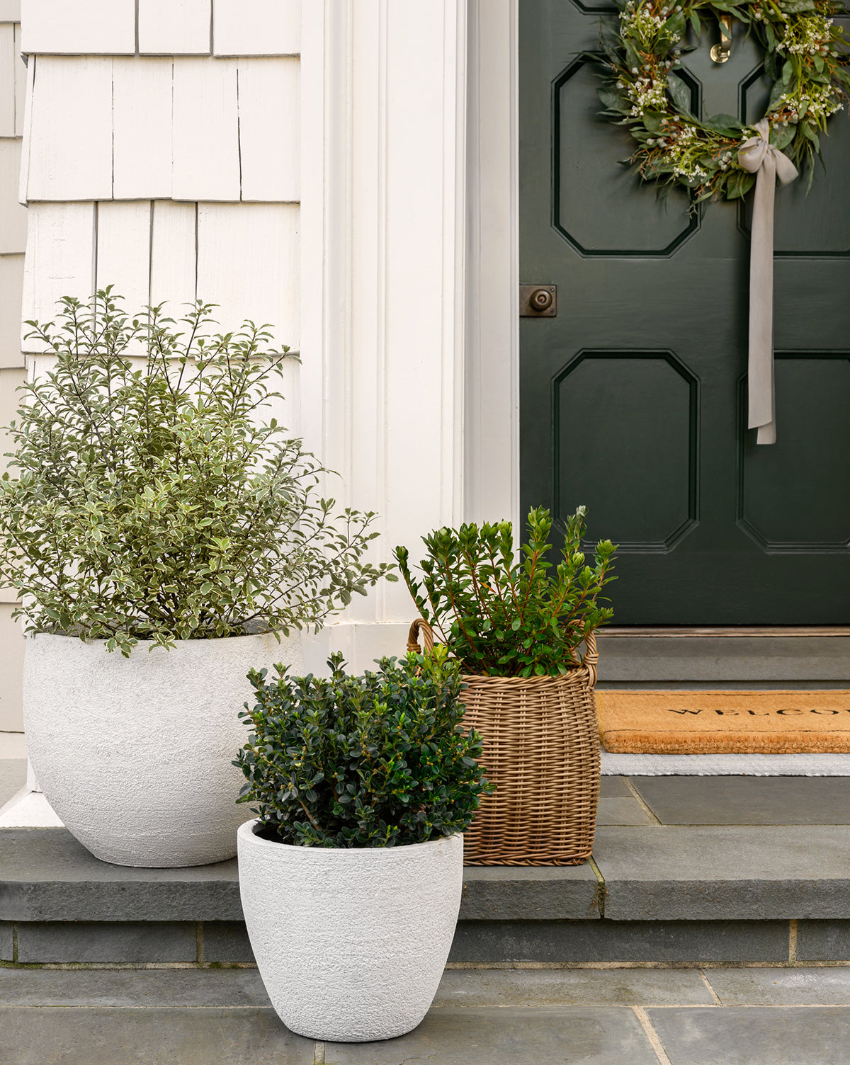 Three potted plants—two in white Bodie Fiberstone Planters and one in a woven basket—sit on stone steps by a dark green front door with a wreath and doormat, beside a light-colored exterior wall.