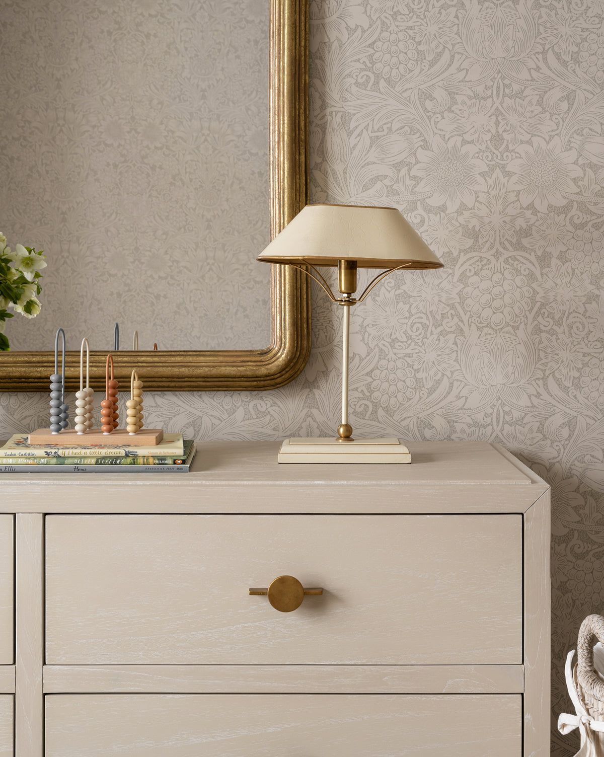 A beige dresser with a round gold knob stands against patterned wallpaper. On top, the Daphne Table Lamp with a beige shade sits next to a gold mirror, books, an abacus, art decor lamp, and a small green plant by McGee & Co.