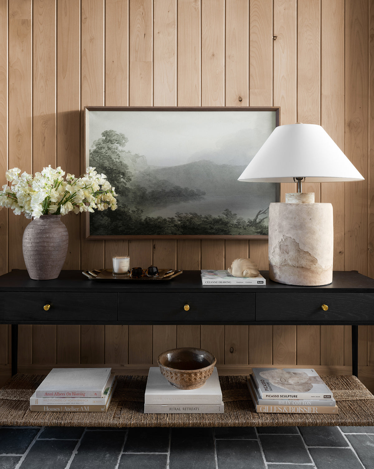 The Dana Console with brass hardware stands against wood paneling, displaying a stone lamp, white florals, a decorative bowl, books, candle, and accents. Misty landscape artwork completes this elegant entryway vignette by McGee & Co.