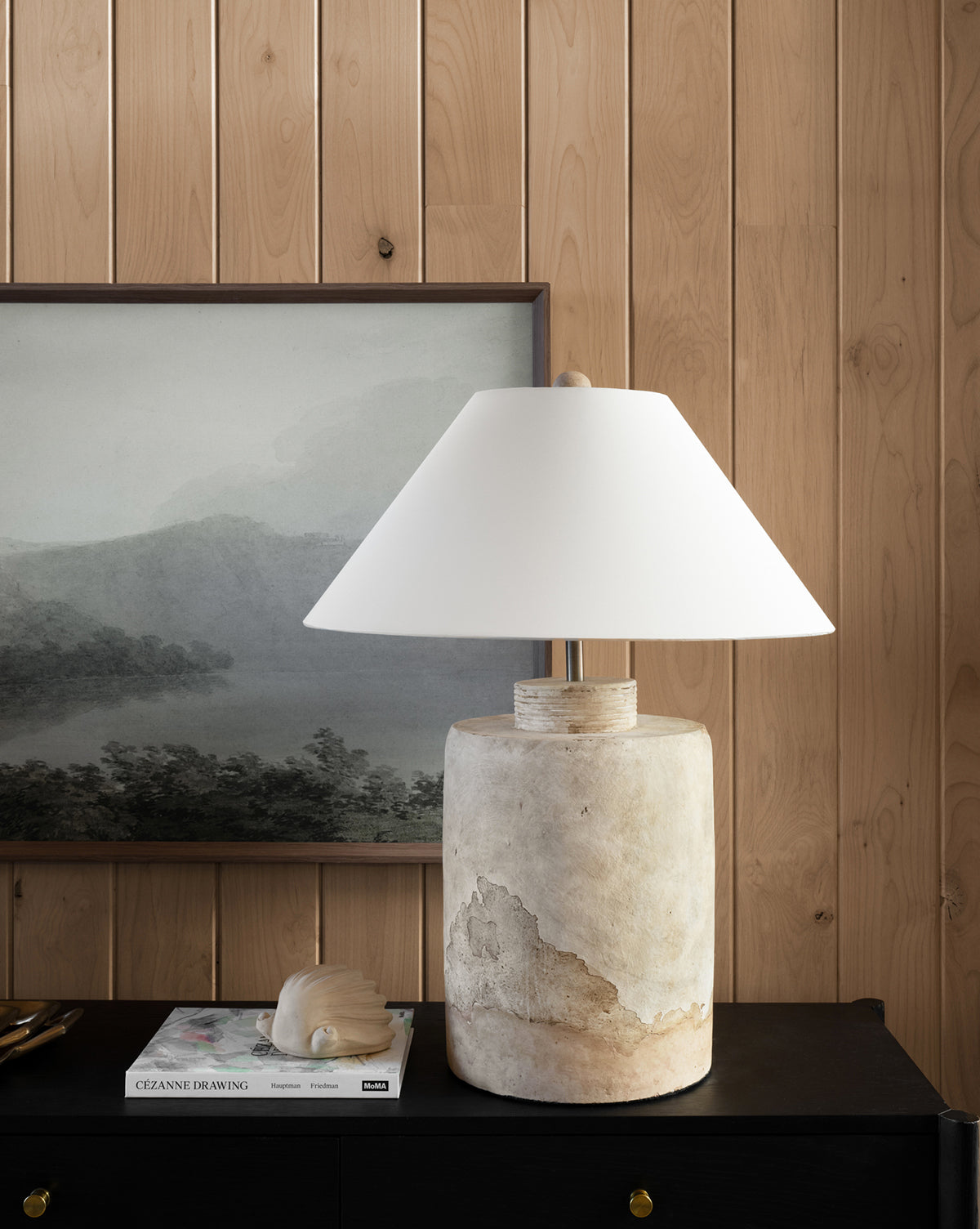 A stone table lamp with a white shade sits on a black cabinet beside a book and the Baroque Shell Object, set against a wood-paneled wall and a muted landscape painting.