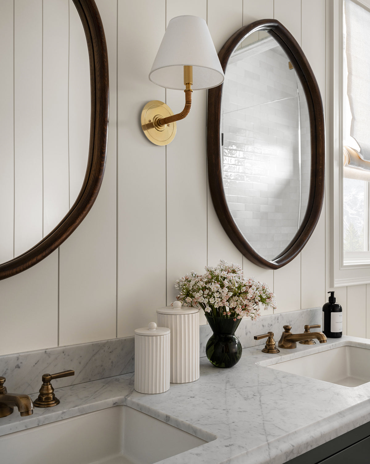 A bathroom vanity with a marble countertop, dual sinks and gold fixtures, two oval mirrors, a wall sconce with white shade, a Fluted Lidded Bathroom Canister, a vase of flowers, plus a window dressed in a roman shade.