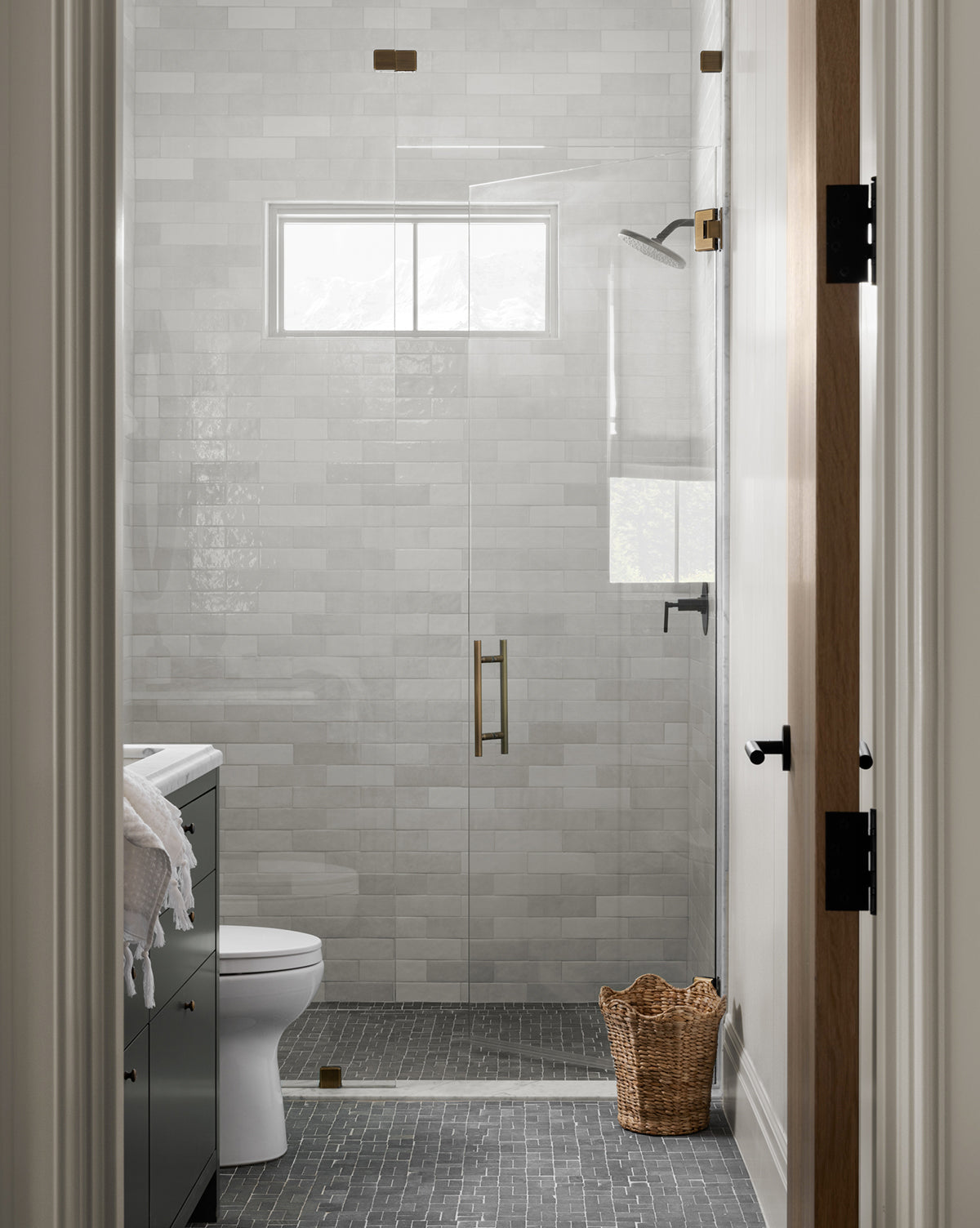 A modern bathroom features light gray subway tile walls, a glass shower, white toilet, dark vanity with towel, gray floor tiles, small window, and the Tenley Woven Waste Bin on the floor.