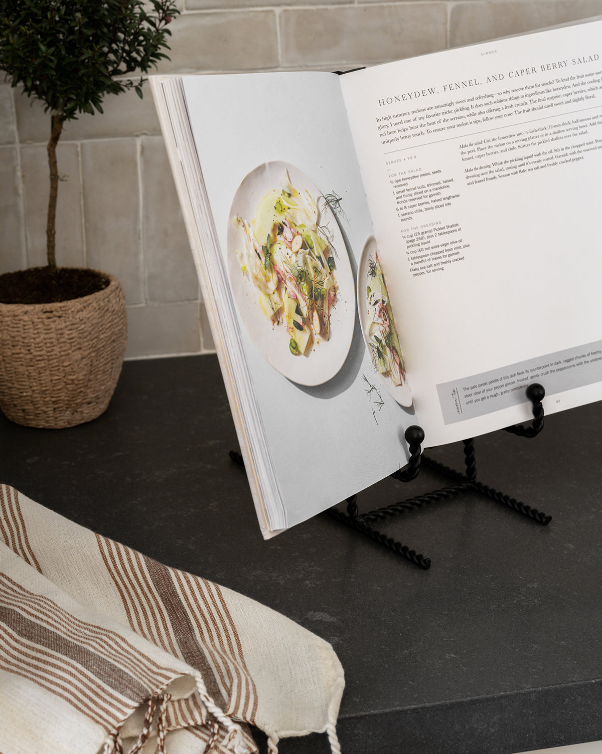 The Finlay Cookbook Holder displays an open cookbook with a salad recipe and plate images on a dark kitchen countertop, styled effortlessly with a potted plant and striped dish towel.