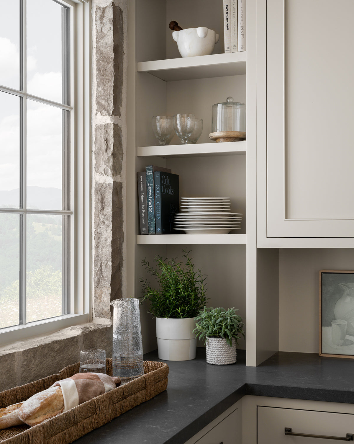 A corner kitchen shelf with dishes, glasses, cookbooks, and plants sits by a window with a view of green hills. On the counter is a Hand-Woven Bankuan Tray holding a loaf of bread and glassware, bringing natural texture to the space.