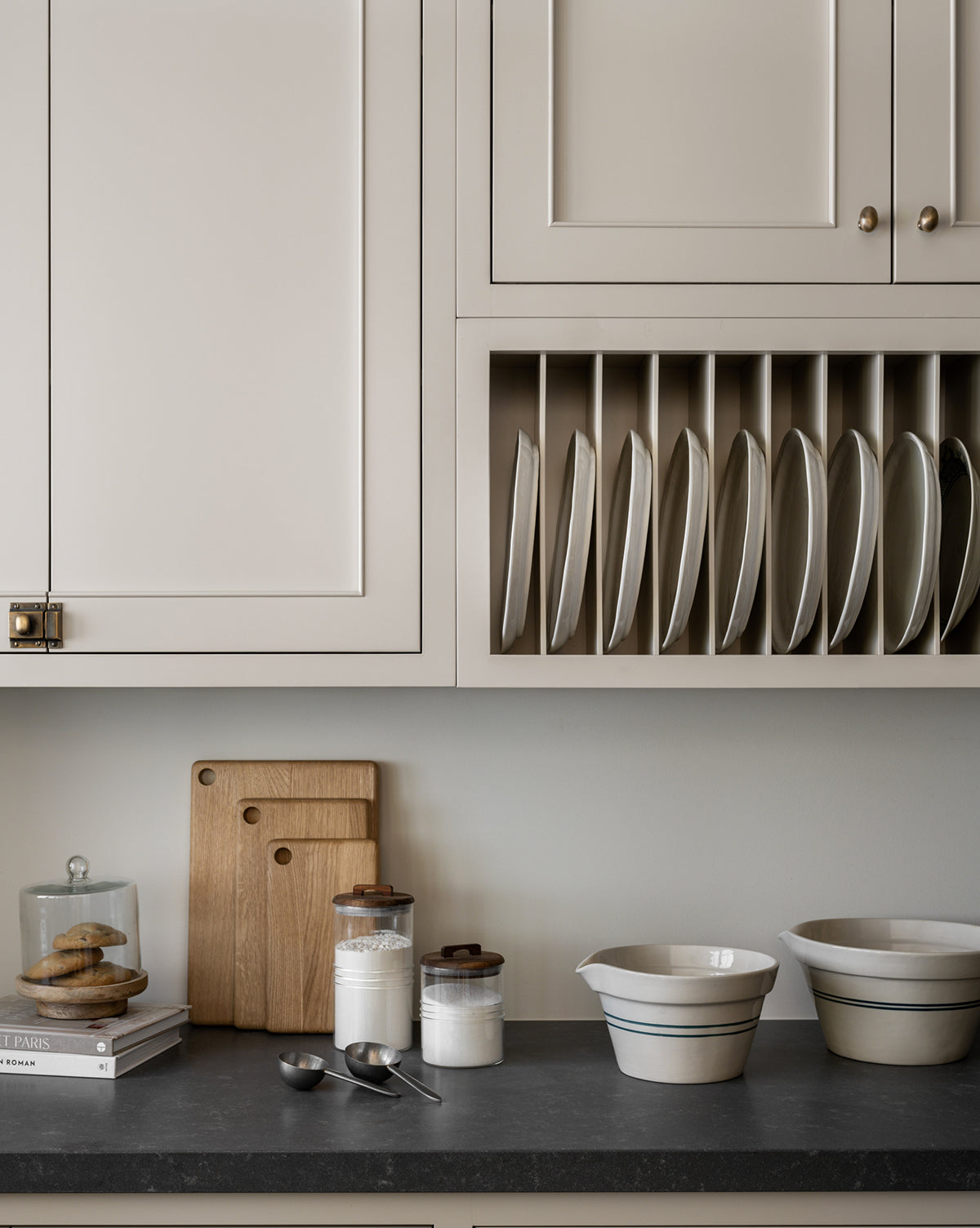 A modern kitchen counter with three wooden cutting boards, a glass jar of cookies, a stack of books, jars of flour and sugar, and an Everett Mixing Bowl, by McGee & Co. Plates are neatly stored vertically in a rack above the counter.