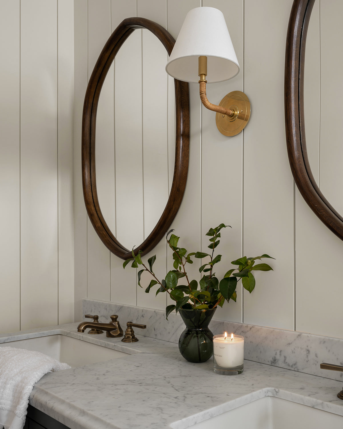 A bathroom vanity with a marble countertop, brass faucet, vase of green branches, lit candle, towel, oval Oslo Mirror with oak frame, and wall-mounted brass sconce with white shade, by McGee & Co.
