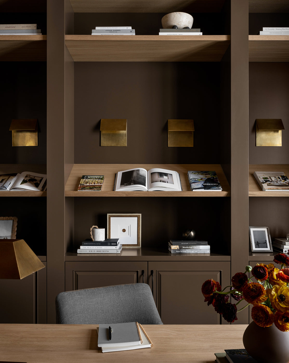 A modern study features Sealed dark brown built-in shelves with books, decor, and brass accents. On the wooden desk, a vase of red and orange flowers stands by an open notebook and pen amid vintage-inspired touches.