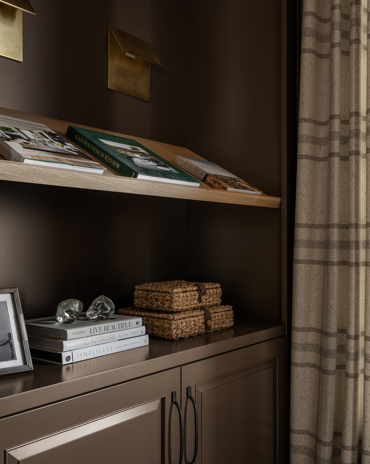 A wooden shelf holding books, magazines, and a Corinne Woven Box sits above a brown cabinet with two black handles; on the cabinet are a framed photo and two glass decor pieces, beside a window dressed in beige plaid curtains.