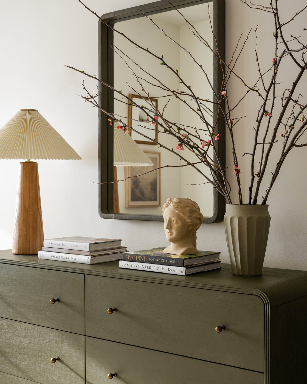 A green dresser holds a stack of books, a beige pleated-shade lamp, a small bust sculpture, and the Frida Vase with branches. Above, a large framed mirror reflects wall art and decor.