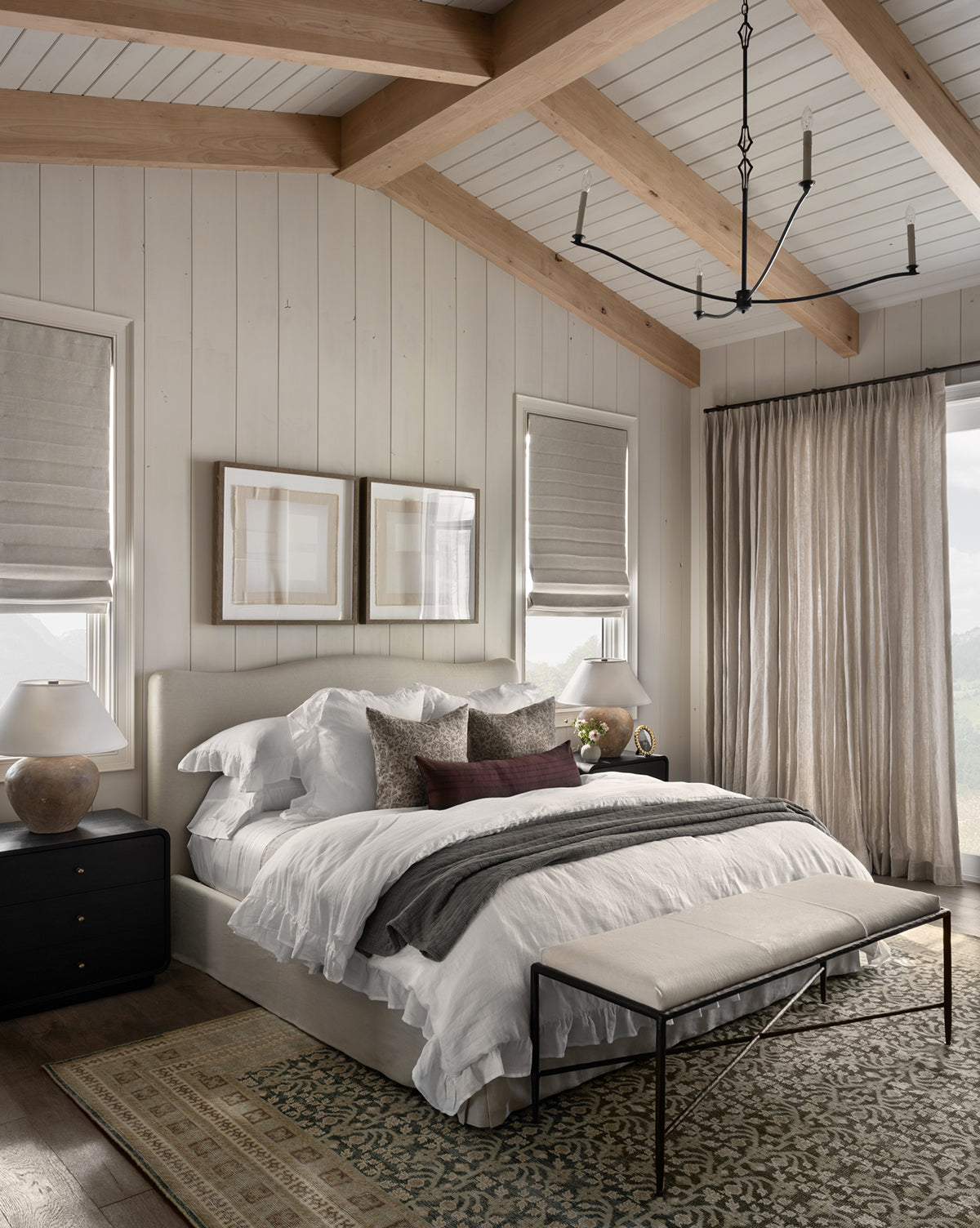 A cozy bedroom with a large bed, white layered bedding, decorative pillows, a foot bench, wood-beamed ceiling, side tables with lamps, two windows with roman shades, and the Evangeline Hand-Knotted Wool Rug for added texture, by McGee & Co.