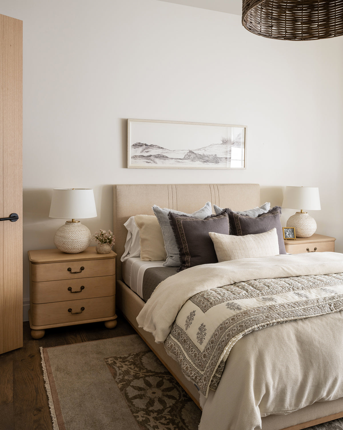 A cozy, neutral-toned bedroom features a neatly made bed, accent pillows, matching wooden nightstands with lamps, and “At the Seashore”—a framed matte sketch—hung above the headboard, all bathed in soft natural light.