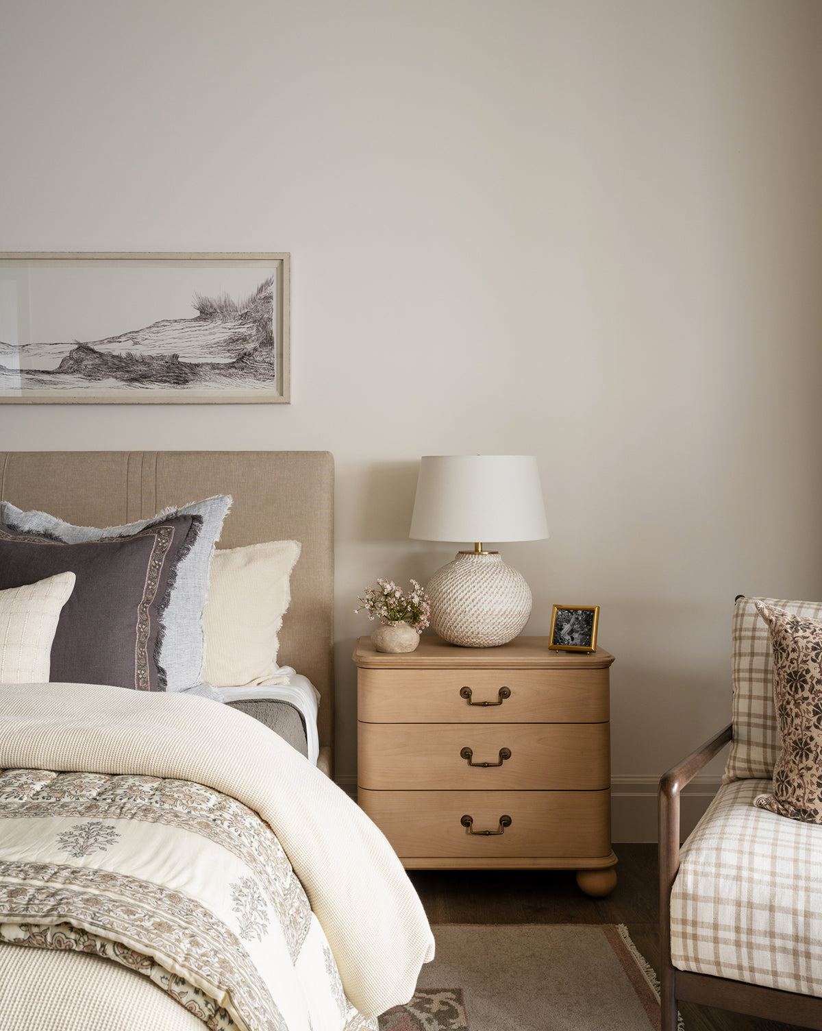 A cozy bedroom with a bed featuring neutral and patterned bedding, a wooden nightstand with an antique brass lamp, small flowers, the Blanche Picture Frame, plus a cushioned chair with plaid pillows against a light wall with art.