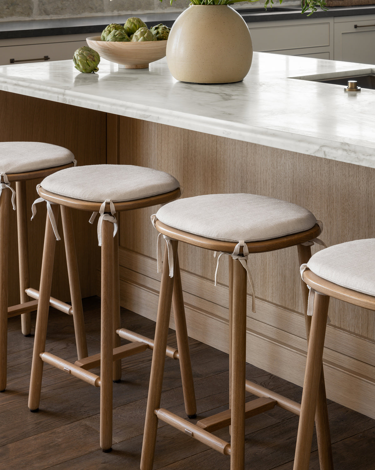 Three Ralph Oak Counter Stools with light linen cushions are lined up along a light wood kitchen counter topped with white marble. A round beige vase and a bowl of artichokes sit on the countertop by McGee & Co.