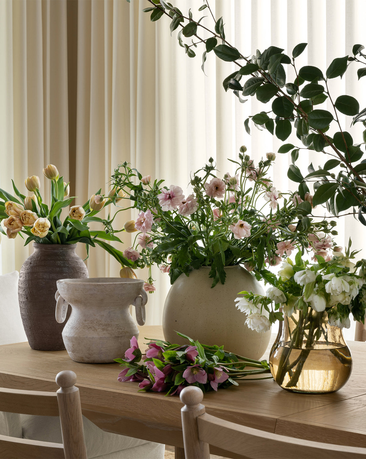 An Arnette Terracotta Vase filled with fresh tulips, daisies, and greenery sits on a wooden table in a softly lit room, cream-colored curtains framing the background.