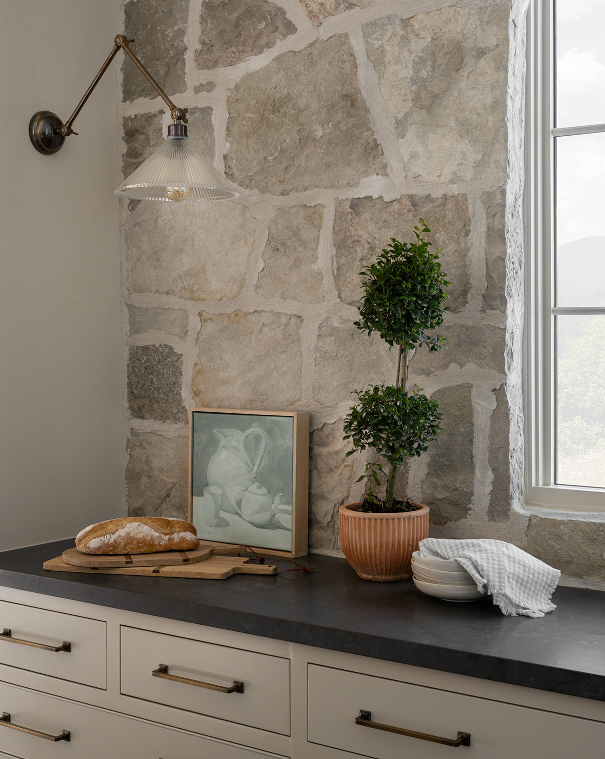 A rustic kitchen features a stone wall, black countertop with a potted plant, the Afternoon Earl Gray giclee art print, a loaf of bread on a wooden board, stacked white bowls with a towel, and a brass wall sconce above.