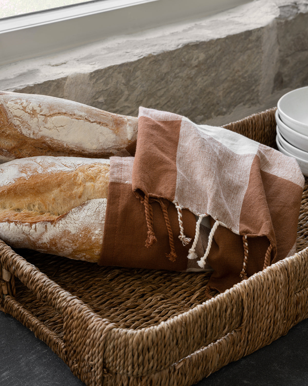 A Hand-Woven Bankuan Tray holds two baguettes wrapped in a checkered cloth, set beside stacked white plates on a stone countertop by the window.