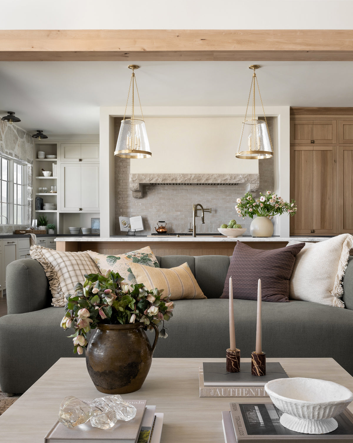 Stylish living room featuring a modern Ines Sofa 90 in gray with neutral pillows, plus a coffee table set with candles, a bowl, and a potted plant. The background highlights a contemporary kitchen with pendant lights and light wood cabinetry, by McGee & Co.