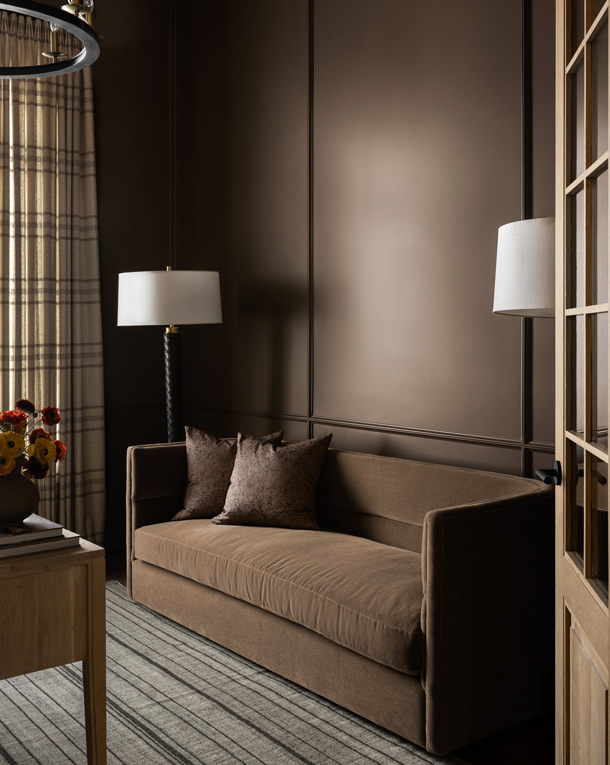 A cozy room with brown walls features a Makerspalm Claremont Sofa 84 with two matching pillows, a striped rug, wooden table with flowers, tall floor lamps with white shades, and plaid curtains over the window—McGee & Co.